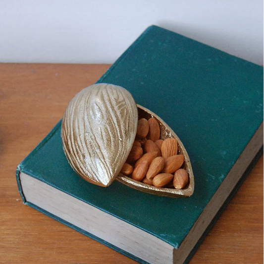 A decorative metallic gold nut-shaped storage box filled with almonds, resting on a dark teal book on a wooden tabletop.
