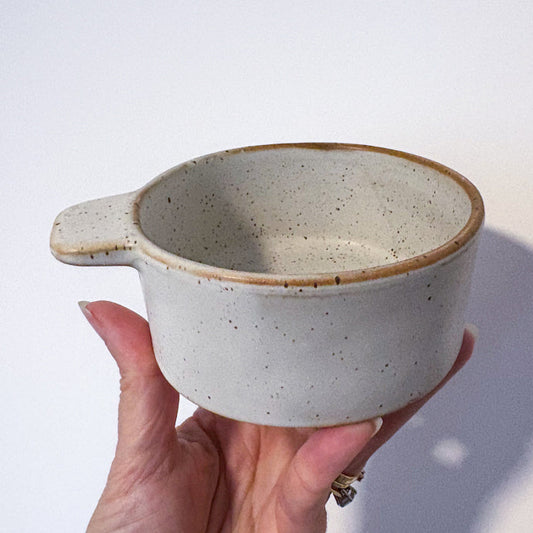 A hand holding a small speckled ceramic cup with a handle, shown against a plain white background with a soft shadow.