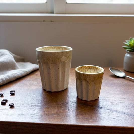 Two handmade ceramic cups with a textured, ribbed flared design and a speckled cream and brown glaze, sitting on a wooden table near a window.