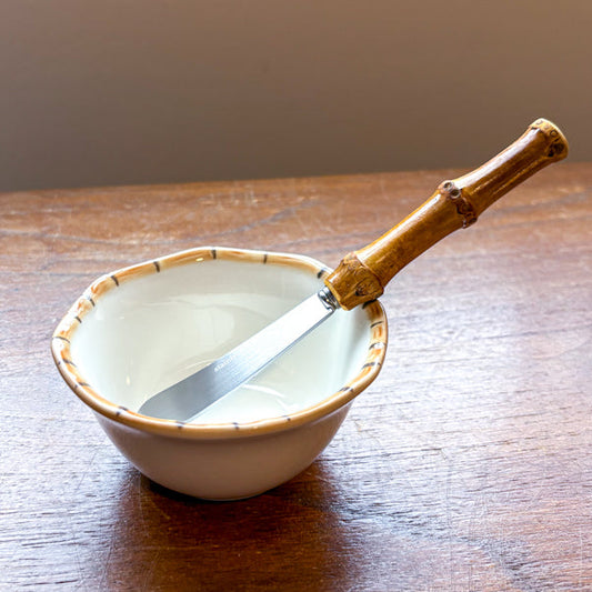 A small ceramic bowl with a bamboo handle metal cheese knife resting inside, placed on a wooden table near a window.