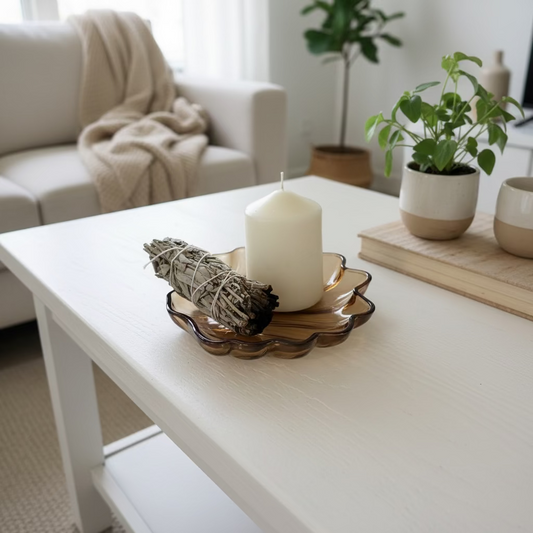 A white pillar candle and a bundled sage smudge rest on a scalloped amber-brown glass dish against a plain off-white wall and tabletop.