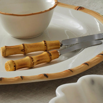 A close-up of a white ceramic table setting with a bamboo-patterned rim, featuring a bowl, knife, and spoon with bamboo handles, arranged on a textured beige cloth.