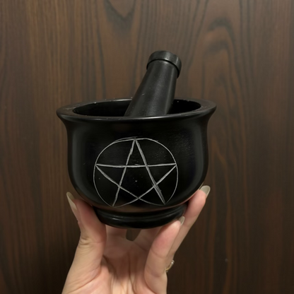 A black stone mortar and pestle with an engraved pentagram symbol on the front, held by a hand against a white paneled background.