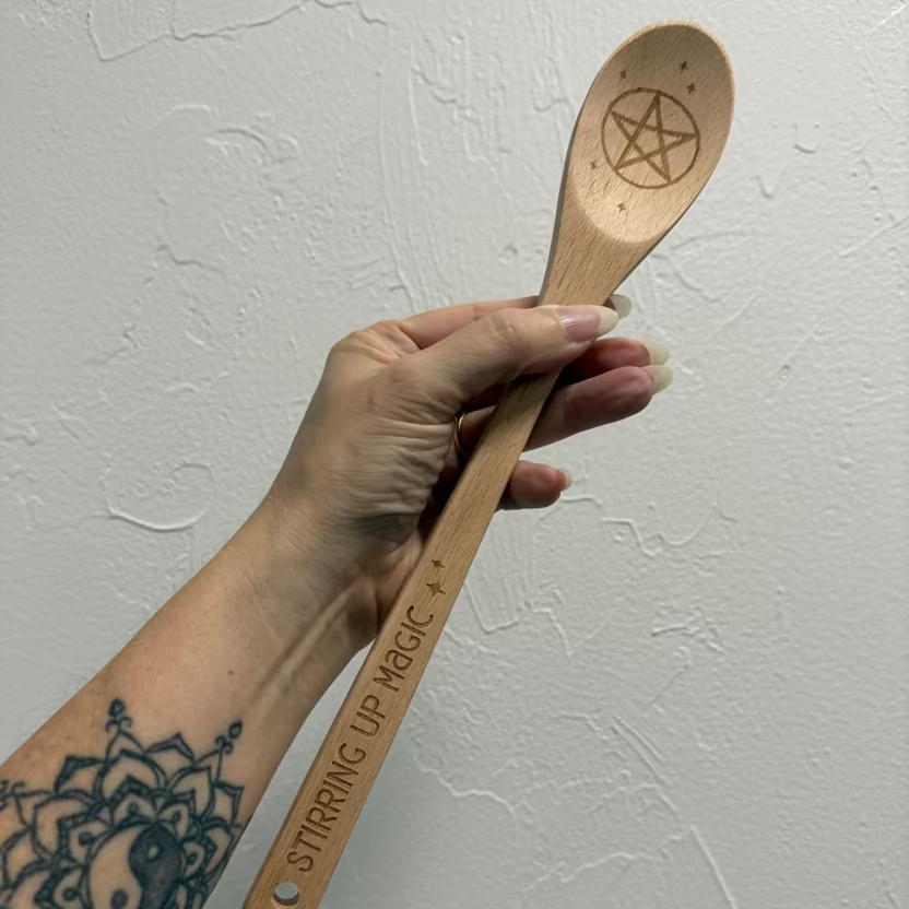 A hand with a mandala tattoo holds a light natural wood spoon against a textured off-white wall. The spoon features a carved pentagram symbol on the bowl and the words 'STIRRING UP MAGIC' printed on the handle.