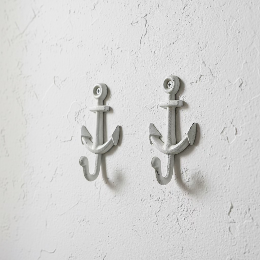 Two white-painted cast iron anchor wall hooks displayed on a wooden cutting board, with a hand pointing to one of the hooks.