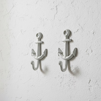Two white-painted cast iron anchor wall hooks displayed on a wooden cutting board, with a hand pointing to one of the hooks.