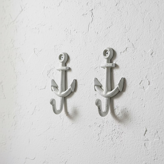 Two white-painted cast iron anchor wall hooks displayed on a wooden cutting board, with a hand pointing to one of the hooks.