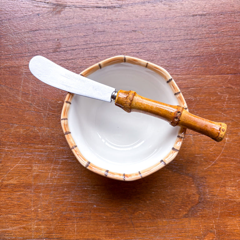 A bamboo butter knife with a stainless steel blade rests on top of a small white bowl with a bamboo-patterned rim, set on a wooden tabletop.