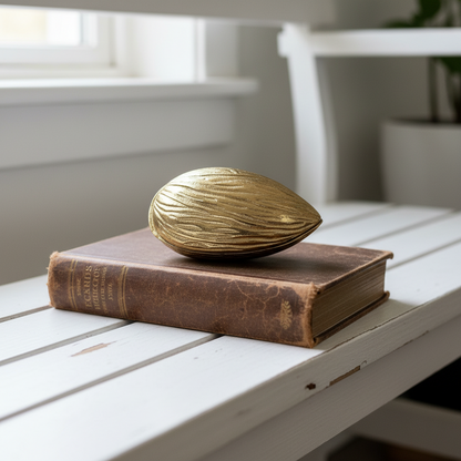 A textured metallic gold bowl with lid resembling a huge nut or seed, resting on top of a vintage brown book on a white wooden slatted table.