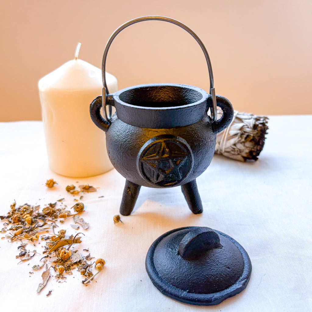 Black cast iron cauldron with pentacle symbol on a white surface with a candle and herbs.