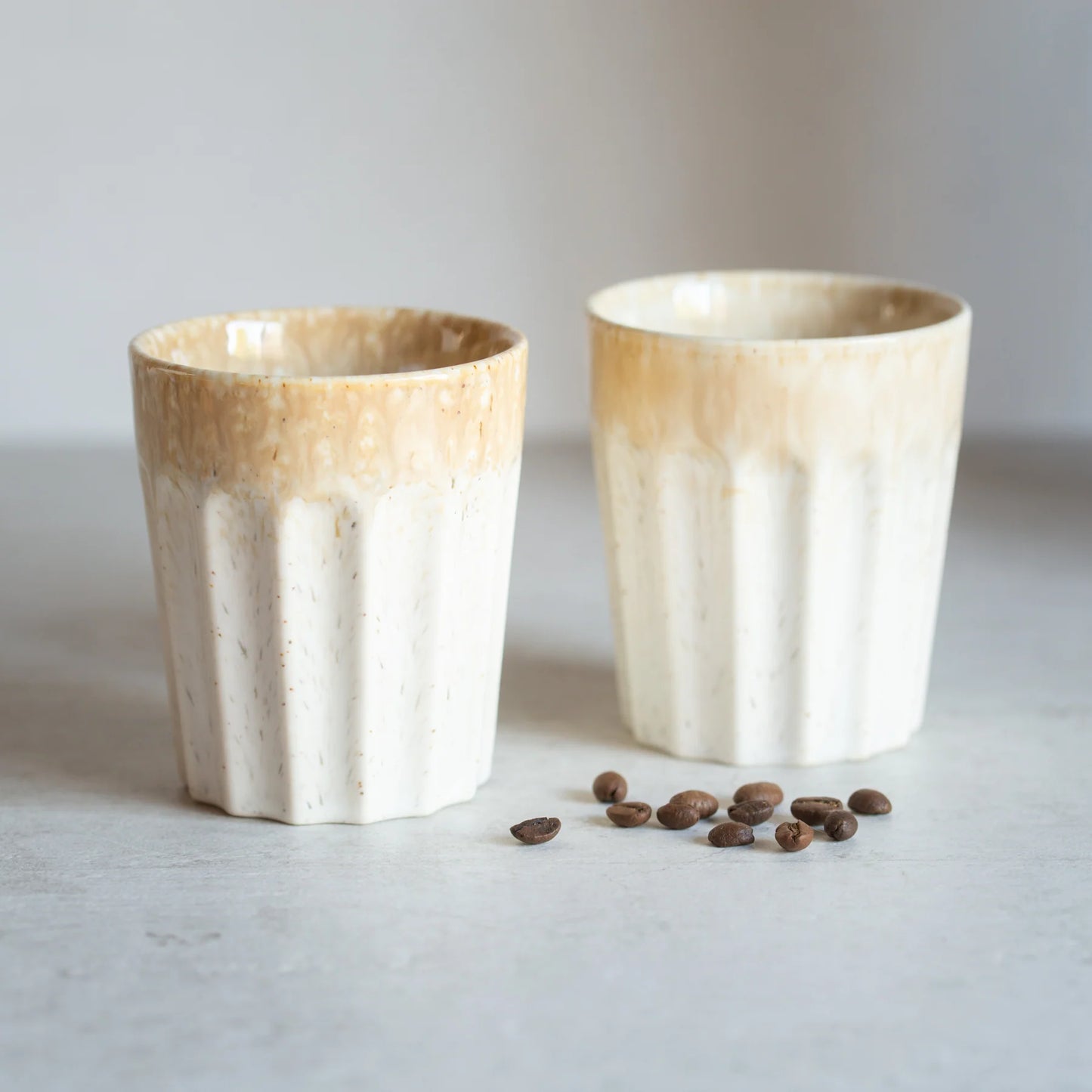Two handmade ceramic tumblers with a fluted design, featuring a cream and beige gradient glaze, displayed on a light gray surface with scattered coffee beans.
