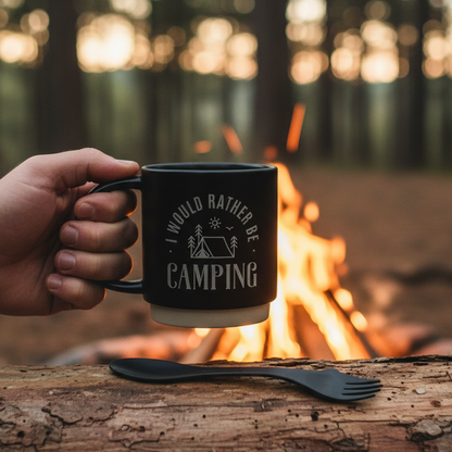 A black ceramic mug with a white printed graphic that reads 'I WOULD RATHER BE CAMPING' with a tent and trees, shown next to a matching black spork