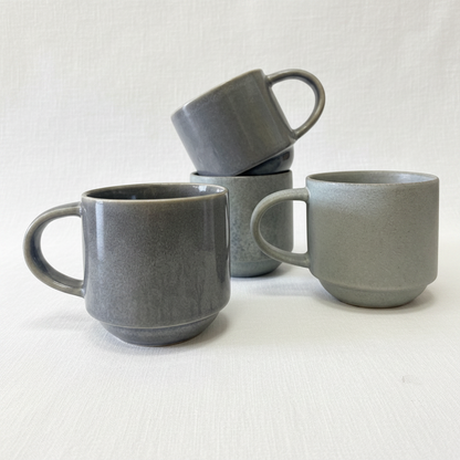 Four matte blue gray ceramic mugs with a textured finish, arranged on a clean white background. One mug in the foreground is upright, another is stacked on top of a third, and a fourth sits to the right.