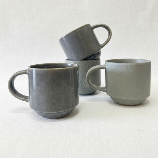 Four matte blue gray ceramic mugs with a textured finish, arranged on a clean white background. One mug in the foreground is upright, another is stacked on top of a third, and a fourth sits to the right.