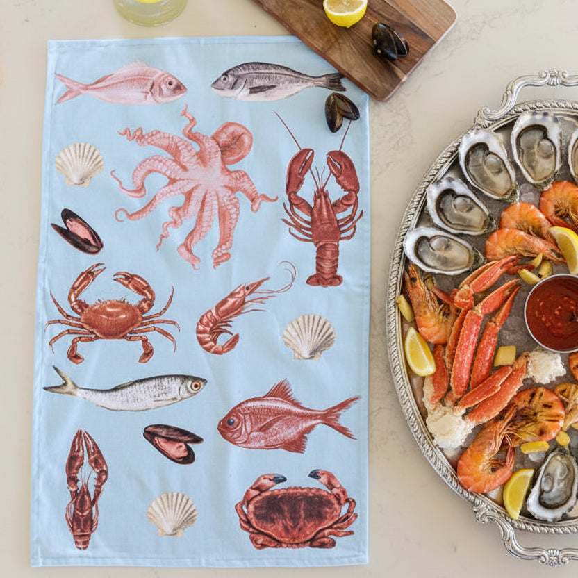 Tea towel with seafood illustrations on a light blue background, placed on a surface with a cutting board and lemon slices.